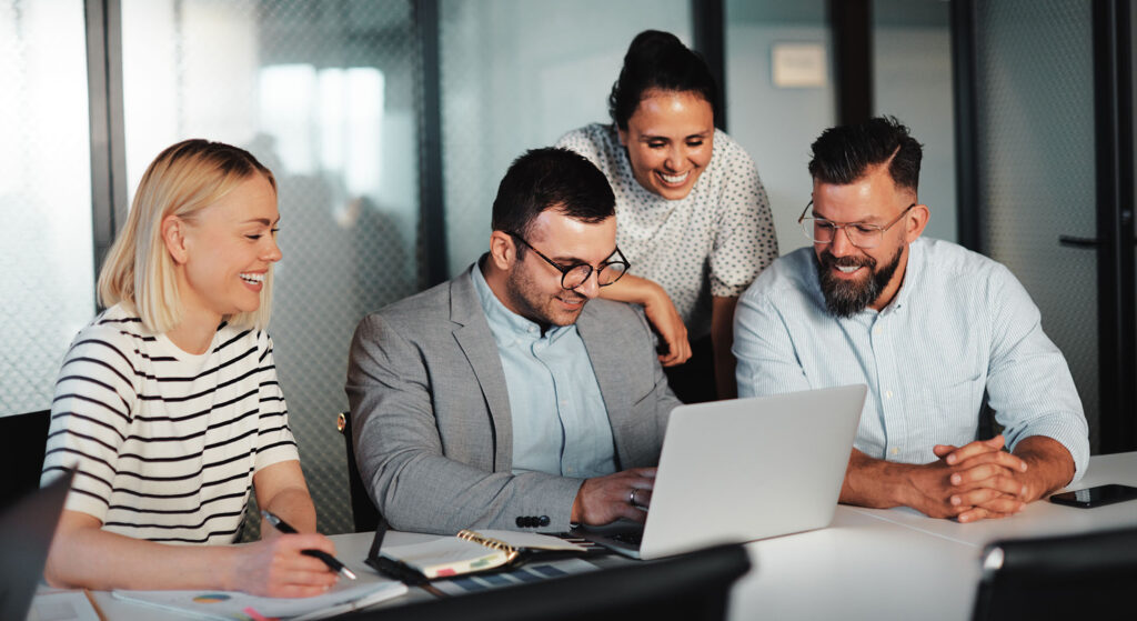 Smiling group of businesspeople working together on a laptop at a table during a meeting in an office boardroom