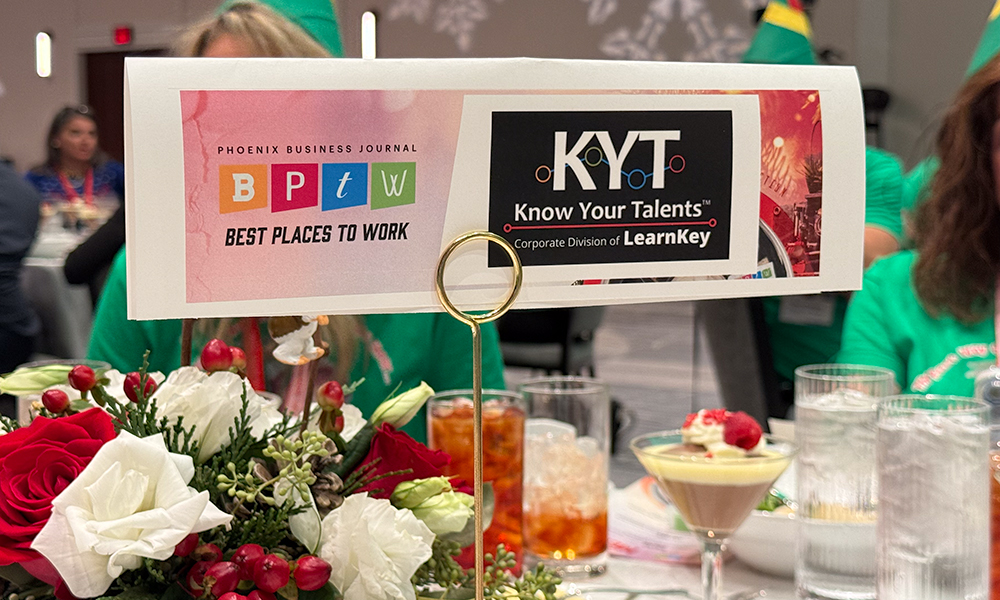 A place card held by a gold holder on a table with drinks and flower centerpiece. The placard reads "Phoenix Business Journal BPTW Best Places to Work: KYT Know Your Talents™ Corporate Division of LearnKey